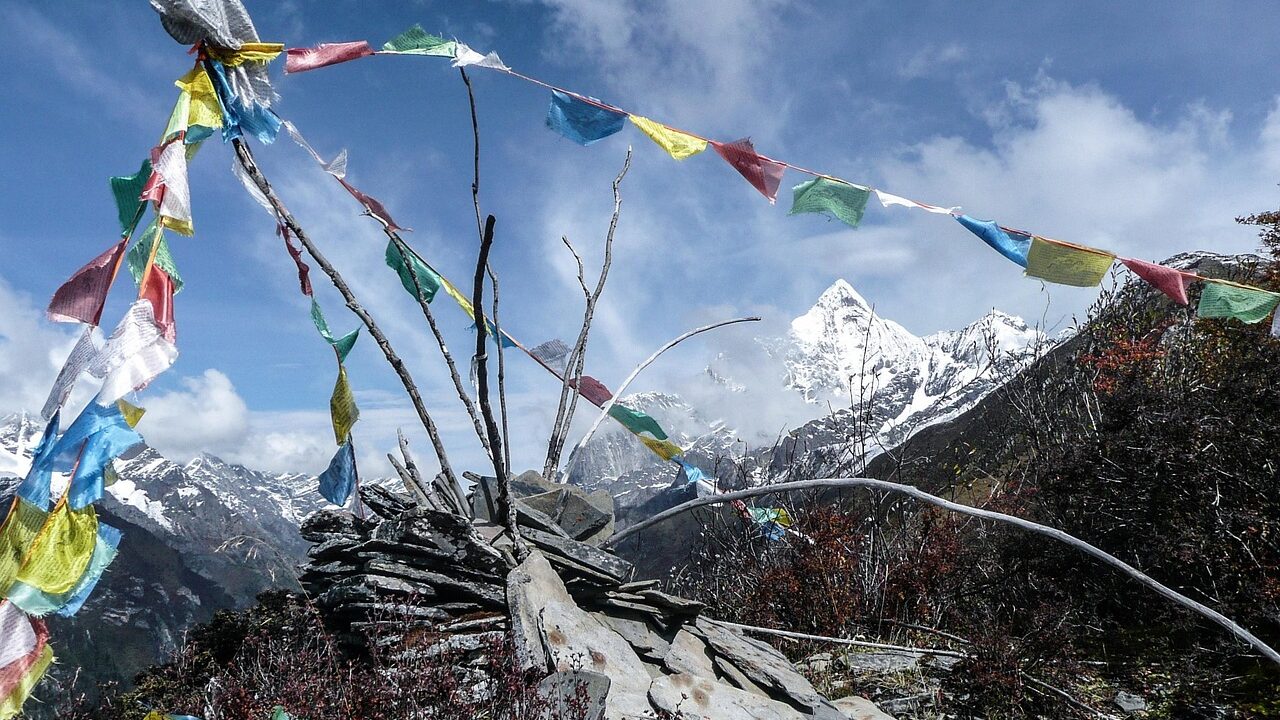 Snow-dusted mountain pass at Kunzum La in Himachal Pradesh, with rugged peaks, a winding dirt road, and prayer flags fluttering in the wind under a partly cloudy sky.