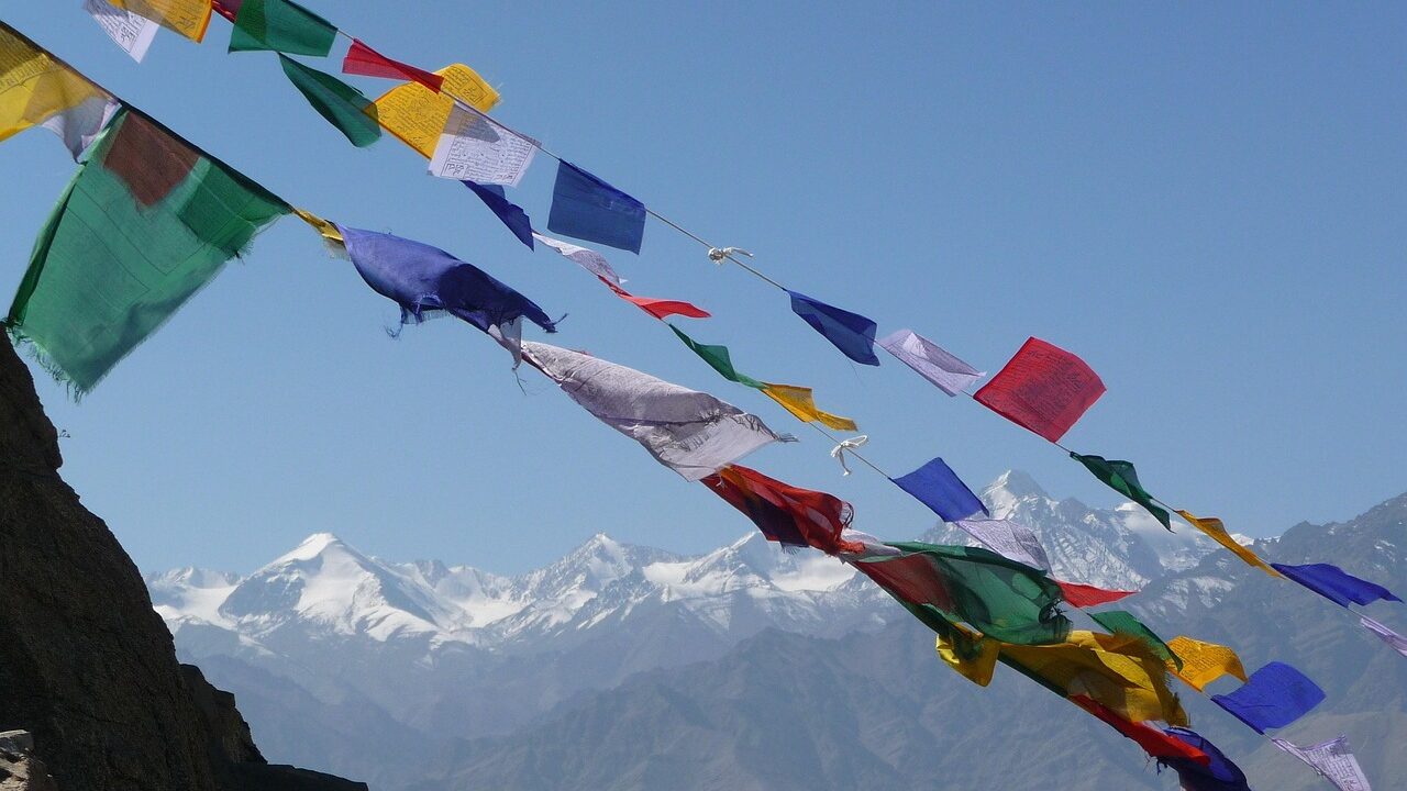 High-altitude mountain pass at Khardung La with a snow-lined road, colorful prayer flags fluttering near a signboard showing the elevation, and rocky Ladakh mountains stretching into the distance.