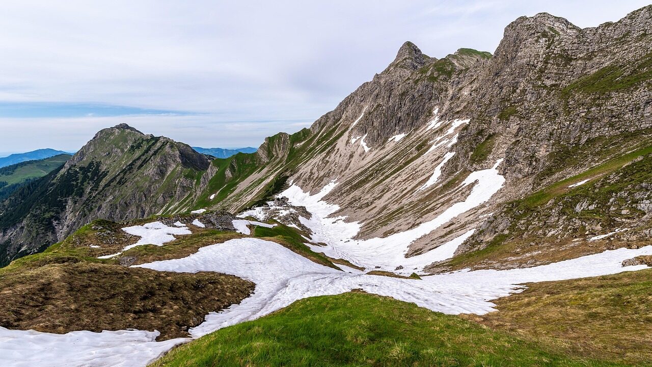 Misty mountain trail leading to Chandrashila Peak, with wildflowers along the path, clouds covering the slopes, and distant peaks partially hidden by fog.